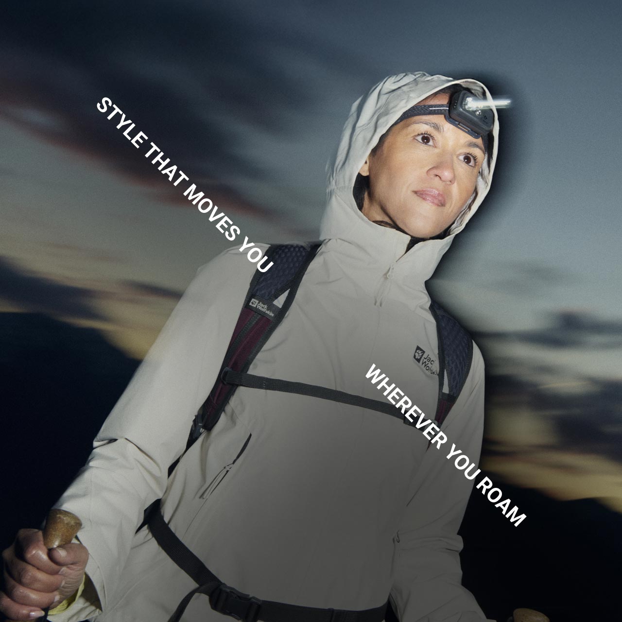 Osoba z outdoorowym wyposażeniem, w tym czołówką, patrzy przed siebie i trzyma kijki trekkingowe o zmierzchu w górach. Tekst brzmi: „Style that moves you wherever you roam”