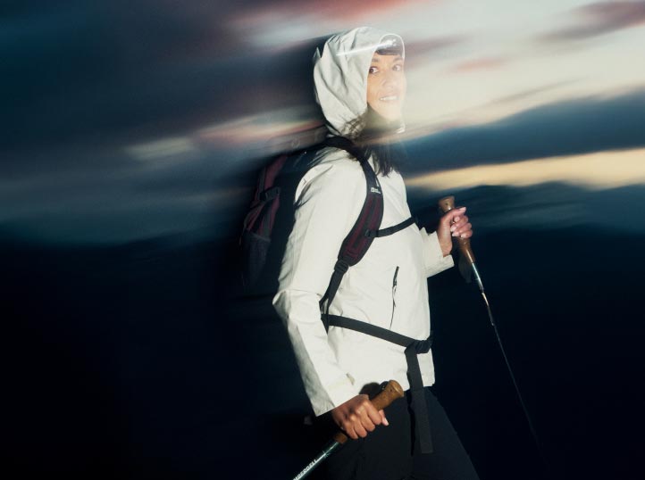 Osoba w odzieży na wędrówki, w tym białej kurtce i kapturze, śmieje się, trzymając w rękach kijki trekkingowe. Rozmyte niebo o zmierzchu przywodzi na myśl ruch i przygodę.
